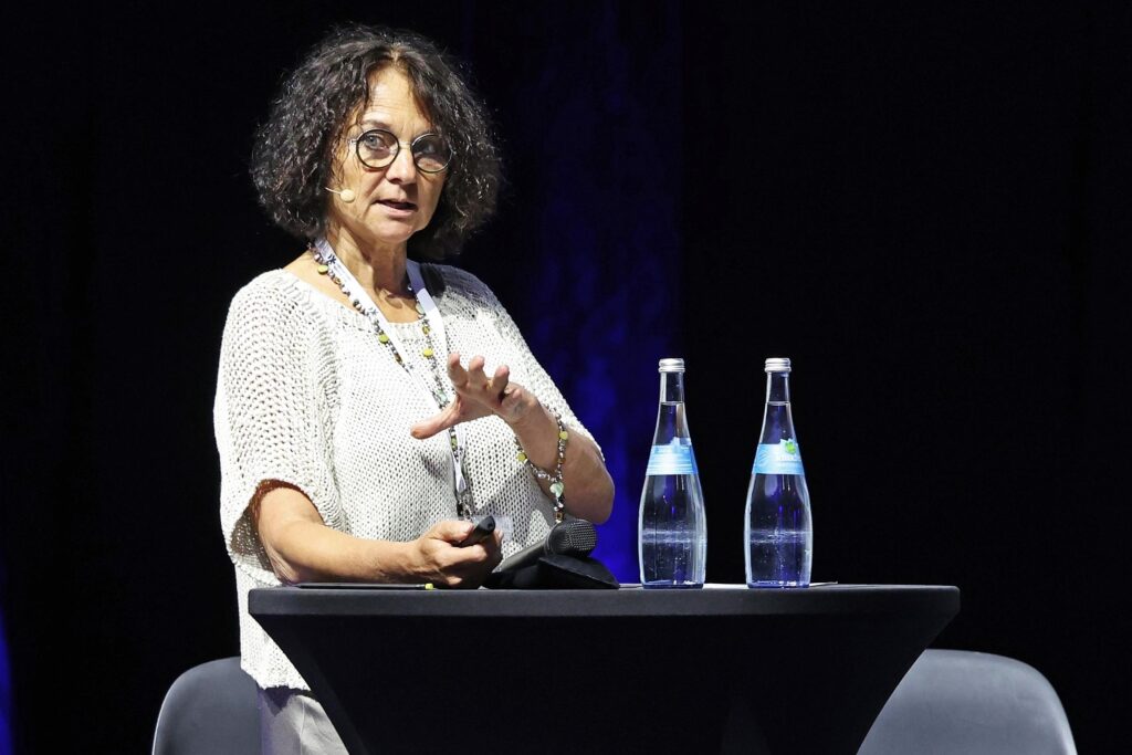 Eine Frau spricht auf einer Konferenzbuehne neben zwei Wasserflaschen.