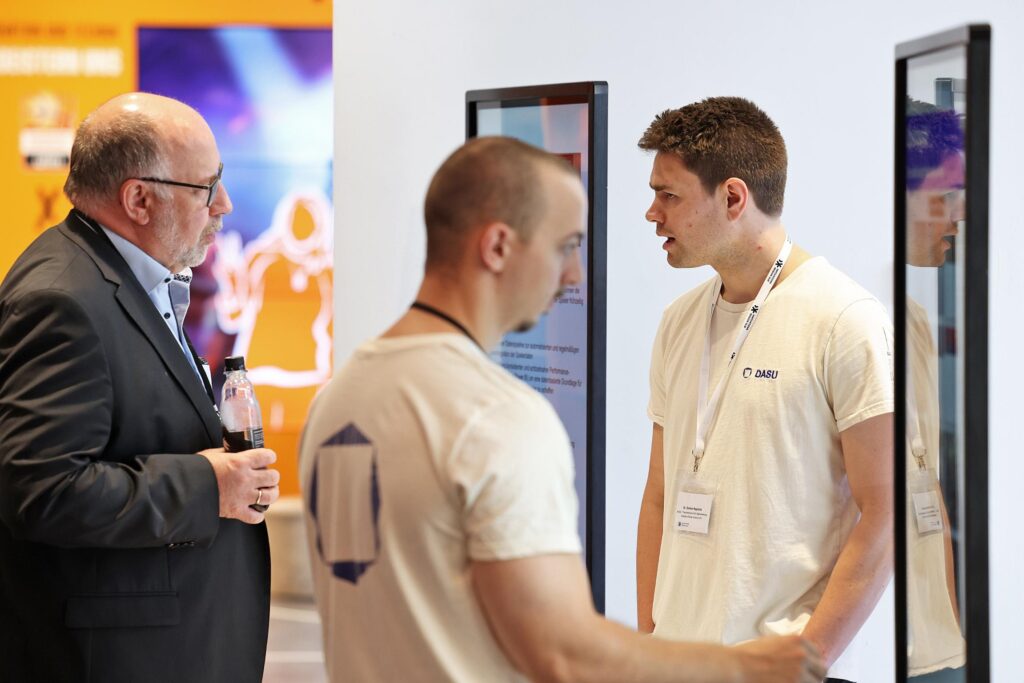 Drei Maenner unterhalten sich vor digitalen Bildschirmen auf einer Messe.