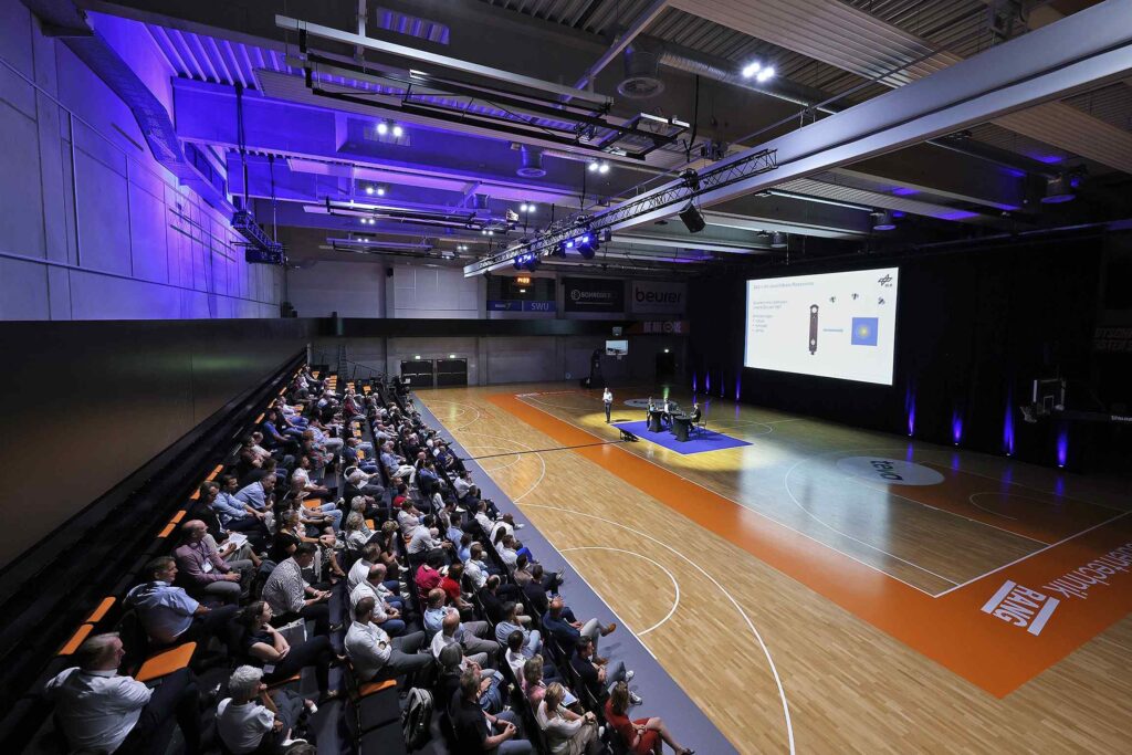 Ein Konferenzraum mit einer großen Leinwand und zahlreichen Teilnehmern.