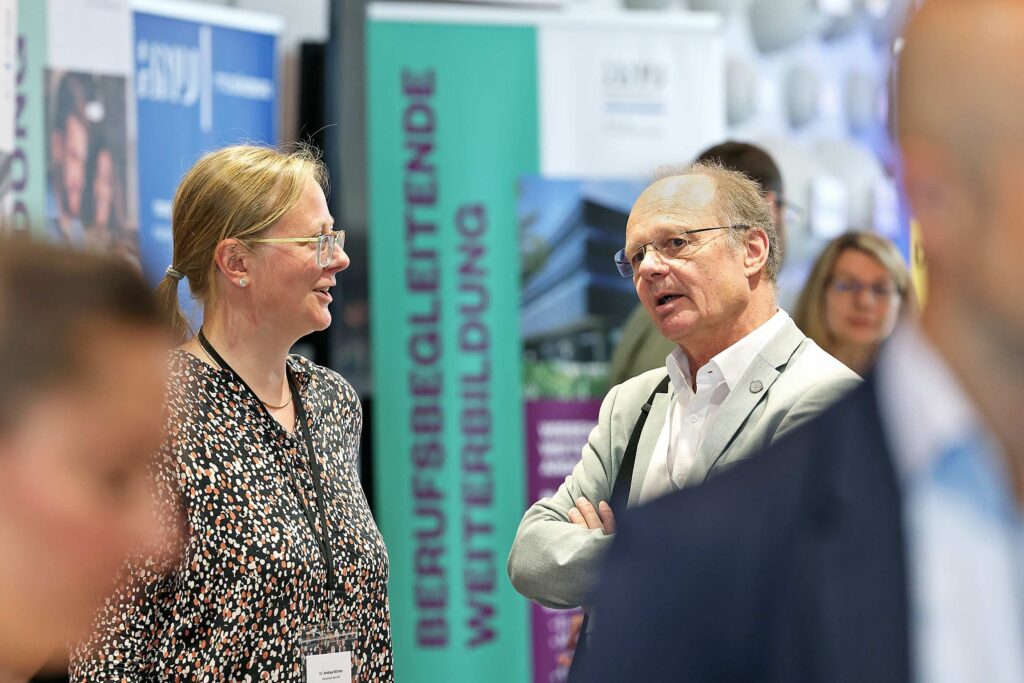 Zwei Personen unterhalten sich auf einer Messe vor einem informativen Hintergrund.