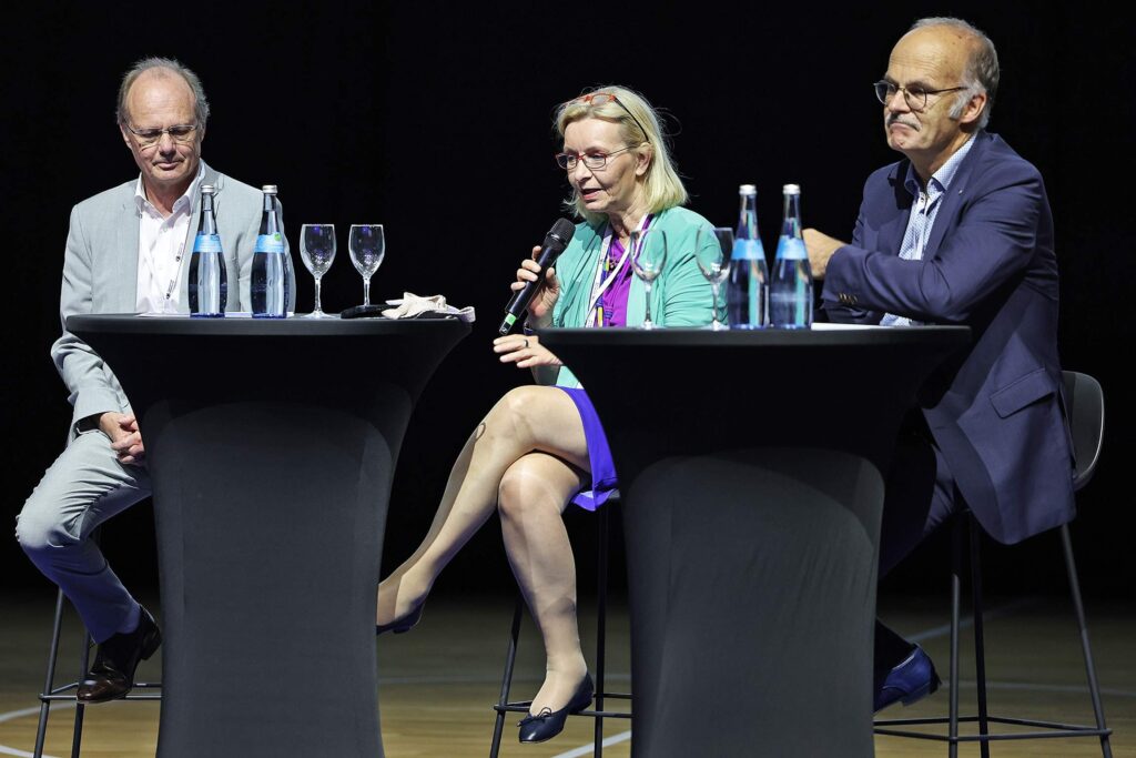 Alt-Text: Drei Personen diskutieren auf einer Buehne bei einer Podiumsdiskussion.